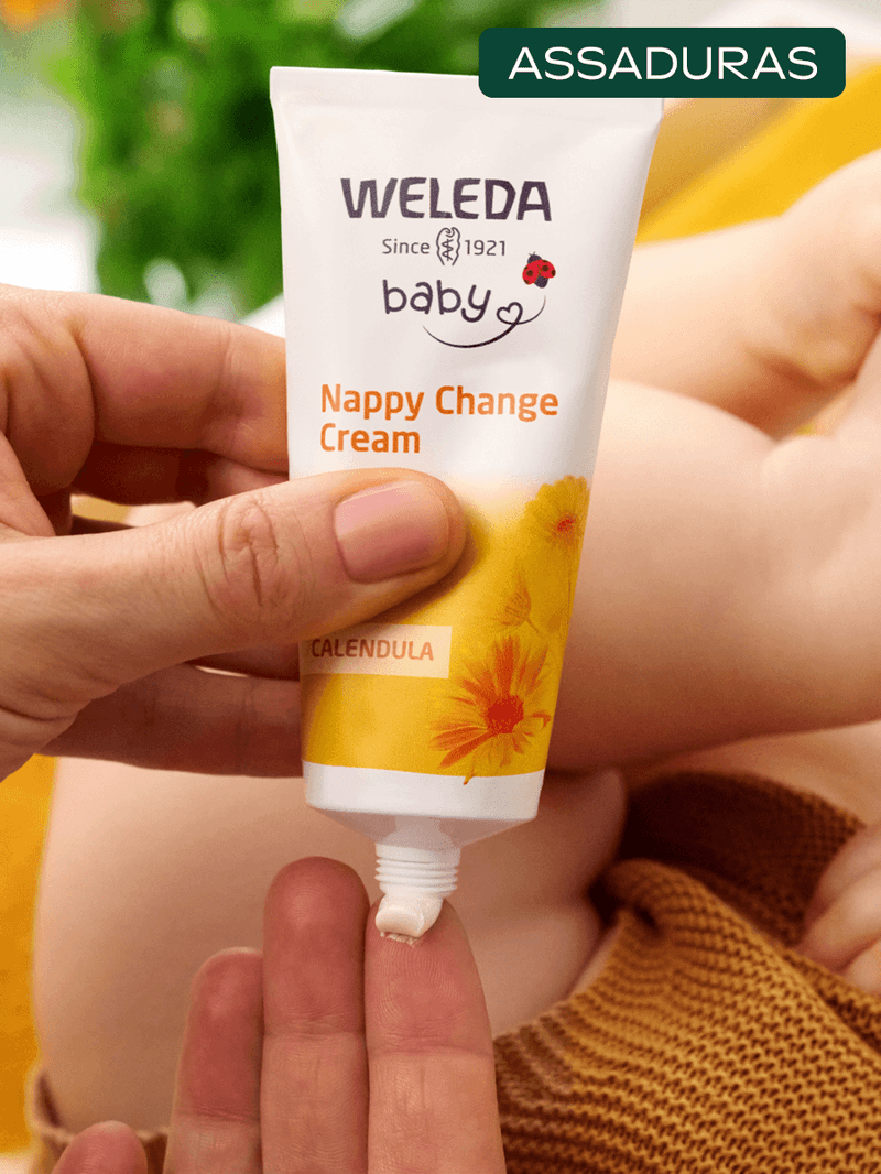 Mao-aplicando-creme-para-assaduras-Weleda-BabyCreme-com-calendula-em-dedo-com-o-produto-visivel-na-embalagem-enquanto-o-bebe-esta-ao-fundo.